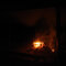 Eine Holzpalette brennt auf einer Feuerstelle. Über ihr und zu einer Seite des Feuers ist es deutlich sichtbar verraucht. Im Vordergrund steht eine in der Dunkelheit kaum zu sehende Holzbank.