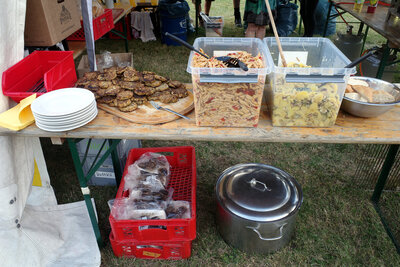 Viel Essen auf einem Festzelttisch auf einer Wiese: Ein Berg von Frikadellen, eine Lagebox mit Nudelsalat, eine Box mit Kartoffelsalat, Weißbrot, unter dem Tisch ein großer Suppentopf und zwei Euroboxen mit weiteren, verpackten Frikadellen. Am Rand des Tischs: Ein Stapel weiter Teller, eine Kiste mit Besteck und eine angebrochene Packung Servietten. Im Hintergrund Kartons, Säcke, Gasflaschen und mehr Tische und Bänke in einem Festzelt. Darauf Getränke und mehr Kisten mit Essen.