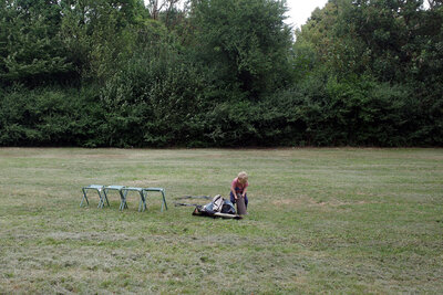 Eine Große, fast leere Wiese vor großem Gebüsch und Bäumen. Alleine in der Mitte des Bildes stehen vier Campingsitze und ein ca. zwei Jahre altes Menschenkind, das mit einer kraftvollen Bewegung den Sack eines noch eingepackten Zeltes auf drückt. Neben ohm liegen ein Stück Zeltstoff und ein kleines Gestänge.
