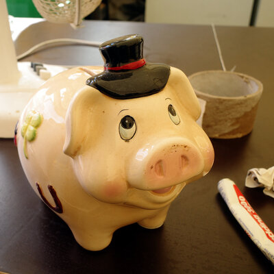Ein farbig bemaltes Keramiksparschwein, das grinst in nach oben schielt. Es trägt einen schwarzen Zylinder und hat ein grünes, vierblättriges Kleeblatt sowie ein Hufeisen auf die rechte Seite gemalt.