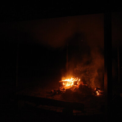 Eine Holzpalette brennt auf einer Feuerstelle. Über ihr und zu einer Seite des Feuers ist es deutlich sichtbar verraucht. Im Vordergrund steht eine in der Dunkelheit kaum zu sehende Holzbank.